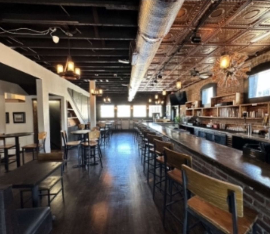 Interior of a bar with a long counter, tables, and seating. Wooden floors, brick, and dark ceiling.