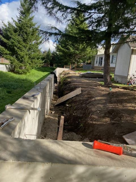 Trenching beside a house foundation with exposed dirt, boards, and a concrete walkway in front