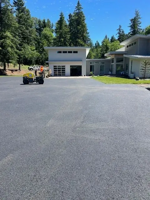 Asphalt driveway in front of a modern white building with garage and transparent door. Trees in background.
