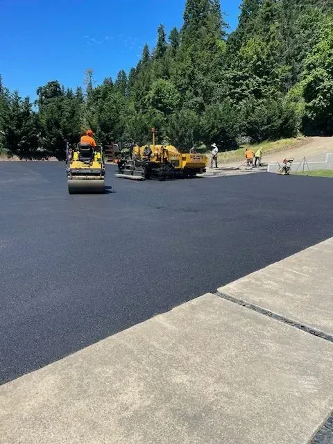 Asphalt paving in progress: Roller compacts fresh black asphalt. Workers and machinery on a sunny day with trees in the background.