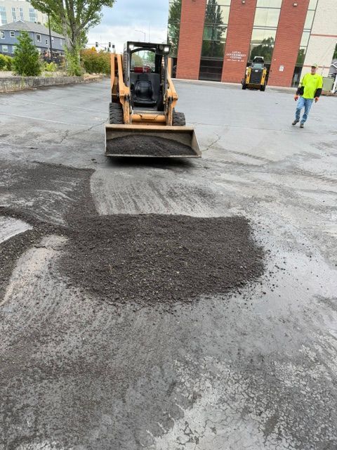 Small construction loader spreading dark asphalt patch on a wet street near a building