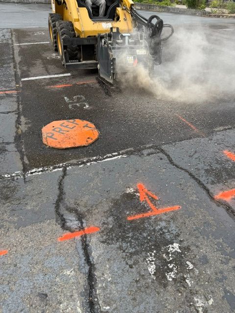 Construction vehicle cutting asphalt, with steam and dust rising on a cracked road marked in orange.