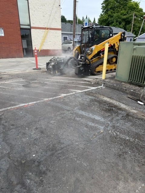 Yellow excavator cutting asphalt near a building, with dust and debris on the pavement.