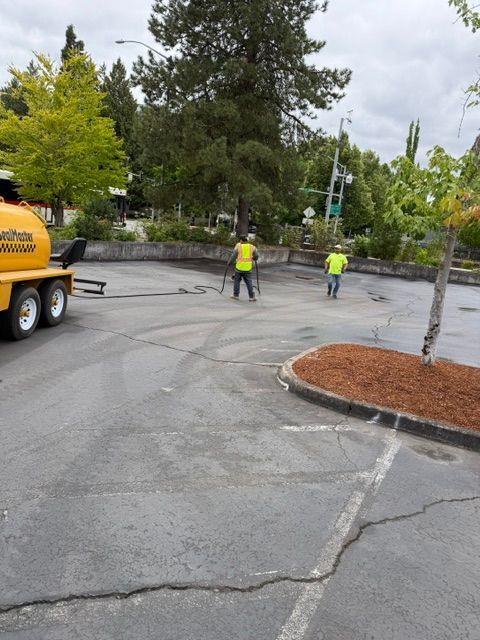 Road crew in neon vests working on a cracked parking lot beside a yellow truck and trees.