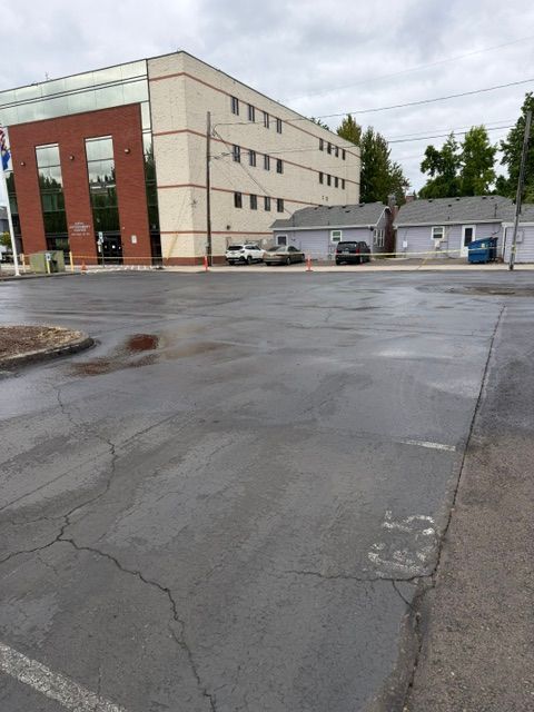 Wet parking lot beside a beige brick building with parked cars and trees under cloudy skies