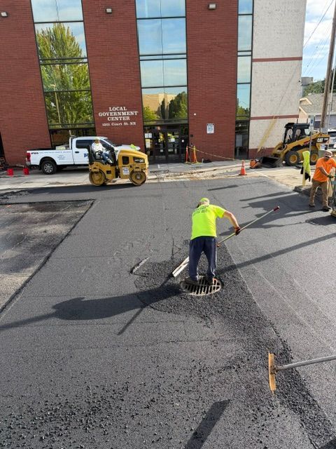 Workers paving asphalt outside a brick building, with a person in a neon vest smoothing fresh pavement.