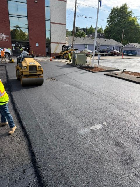 Road resurfacing near a building, with a roller and workers on a wet street.