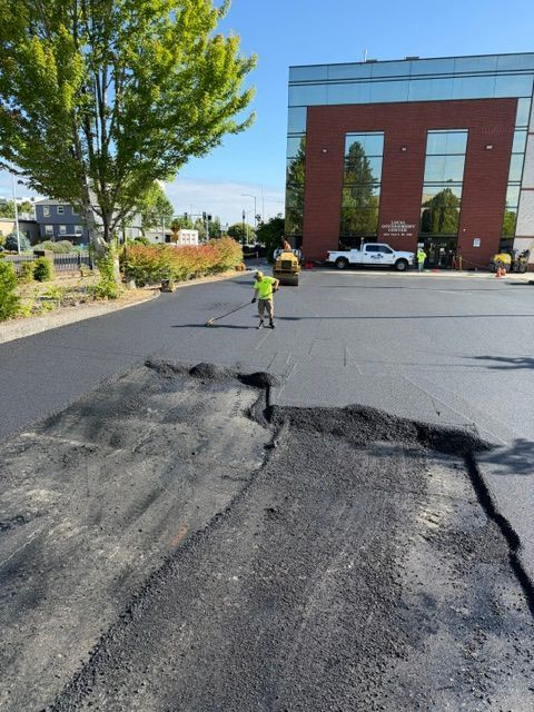 Freshly patched road with cracked asphalt near a brick building; worker in yellow vest ahead