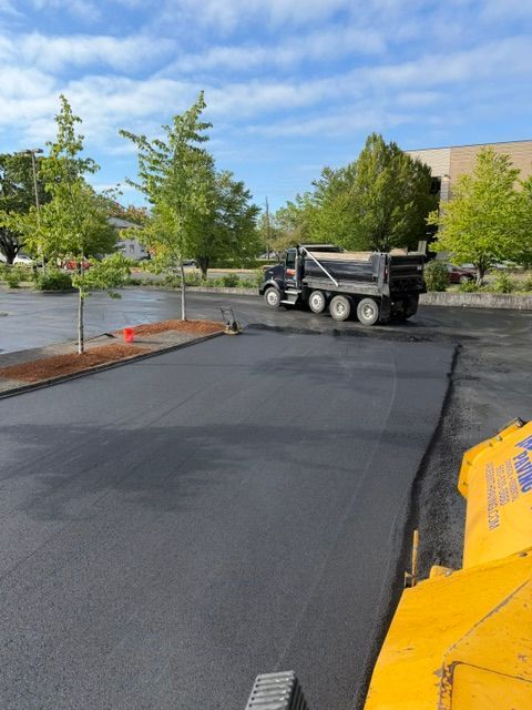 Paving crew smoothing fresh black asphalt in a parking lot beside trees and a building