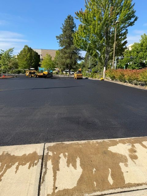 Freshly paved blacktop parking lot with trees, landscaping, and construction equipment in the distance