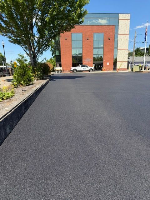 Wide empty asphalt lot beside a red-brick building and trees on a sunny day