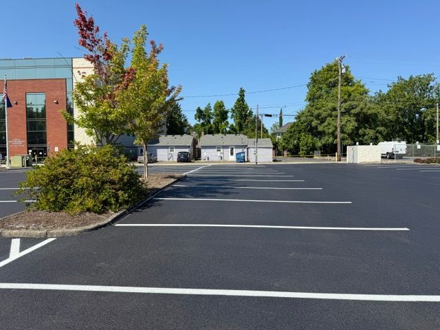 Empty parking lot beside a brick building on a sunny day, with trees and a blue sky.