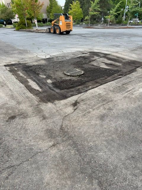 Freshly patched asphalt section on a road, with a small orange construction vehicle in the background.