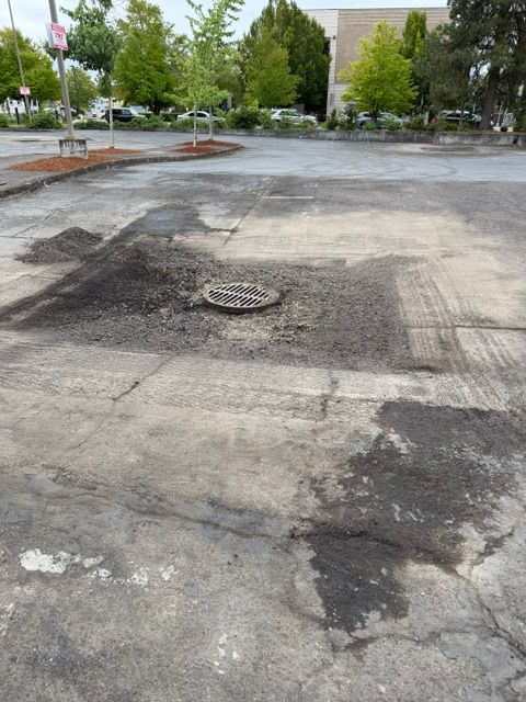 Damaged asphalt parking lot with patches, a manhole cover, and trees in the background