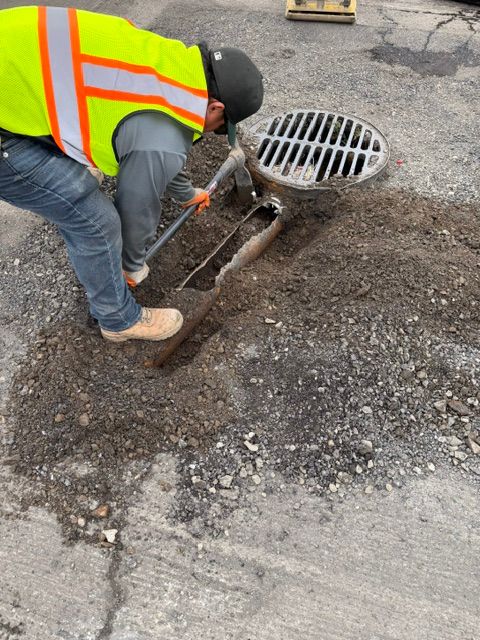 Worker in a neon safety vest clearing debris from a storm drain on a road shoulder