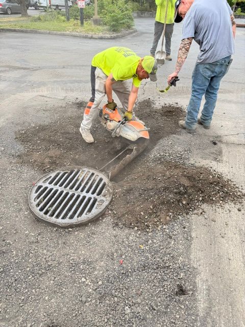Workers repairing potholes on a wet road beside a storm drain