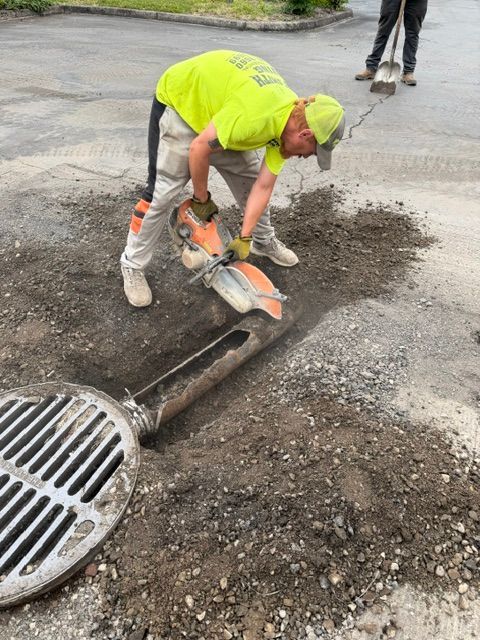 Worker in neon yellow cutting pavement around a storm drain with a concrete saw.