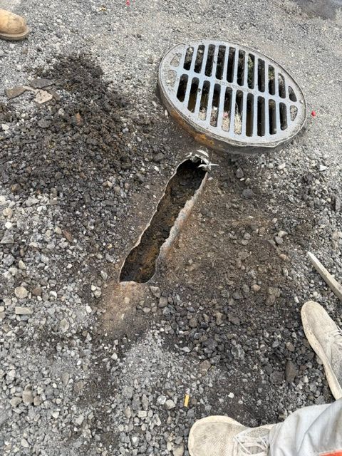 Broken drainage area with a cracked trench beside a round metal storm drain grate on gravel.