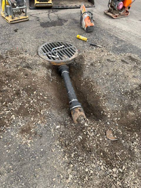 Street drain grate with a black pipe in a construction trench, surrounded by gravel and heavy equipment.