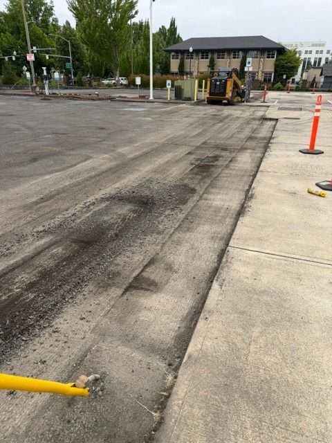 Road construction area with a small excavator, orange cones, and resurfaced pavement near buildings
