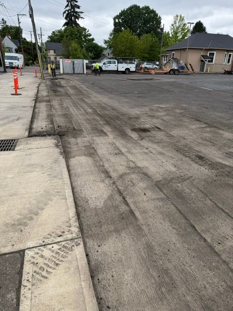 Roadside construction area with fresh asphalt, sidewalk curb, orange cones, and parked vehicles nearby