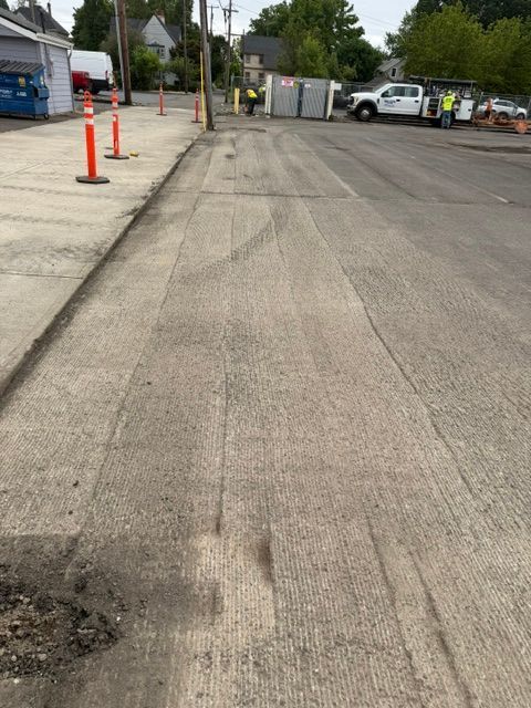 Empty parking lot with an uneven asphalt surface, orange cones, and parked vehicles near a fence.