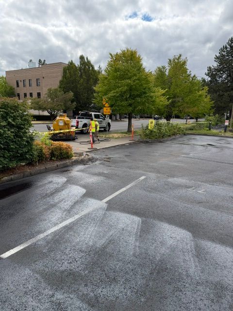 Rainy parking lot with workers and a yellow excavator near trees and a brick building