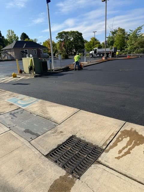 Freshly paved parking lot with workers near a curb and storm drain under clear skies