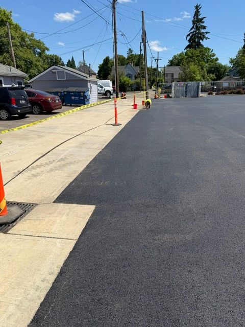 Freshly paved parking lot beside a sidewalk with orange cones and a red car near houses