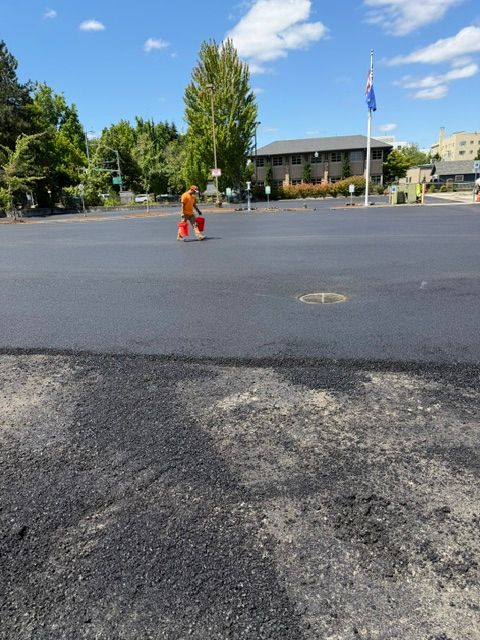 Person in orange stands on a wide paved street near a flagpole and buildings on a sunny day