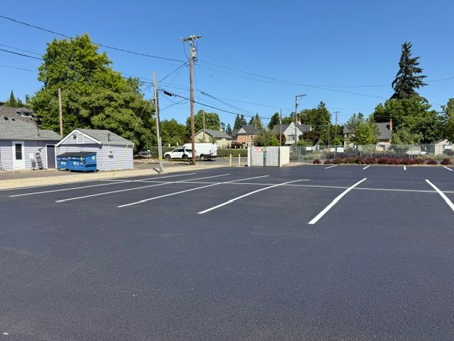 Empty parking lot with white lines, small buildings, utility poles, and trees under a clear blue sky
