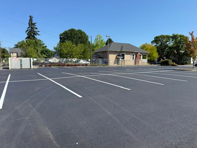 Empty parking lot with white lines beside a small building and trees under a clear blue sky