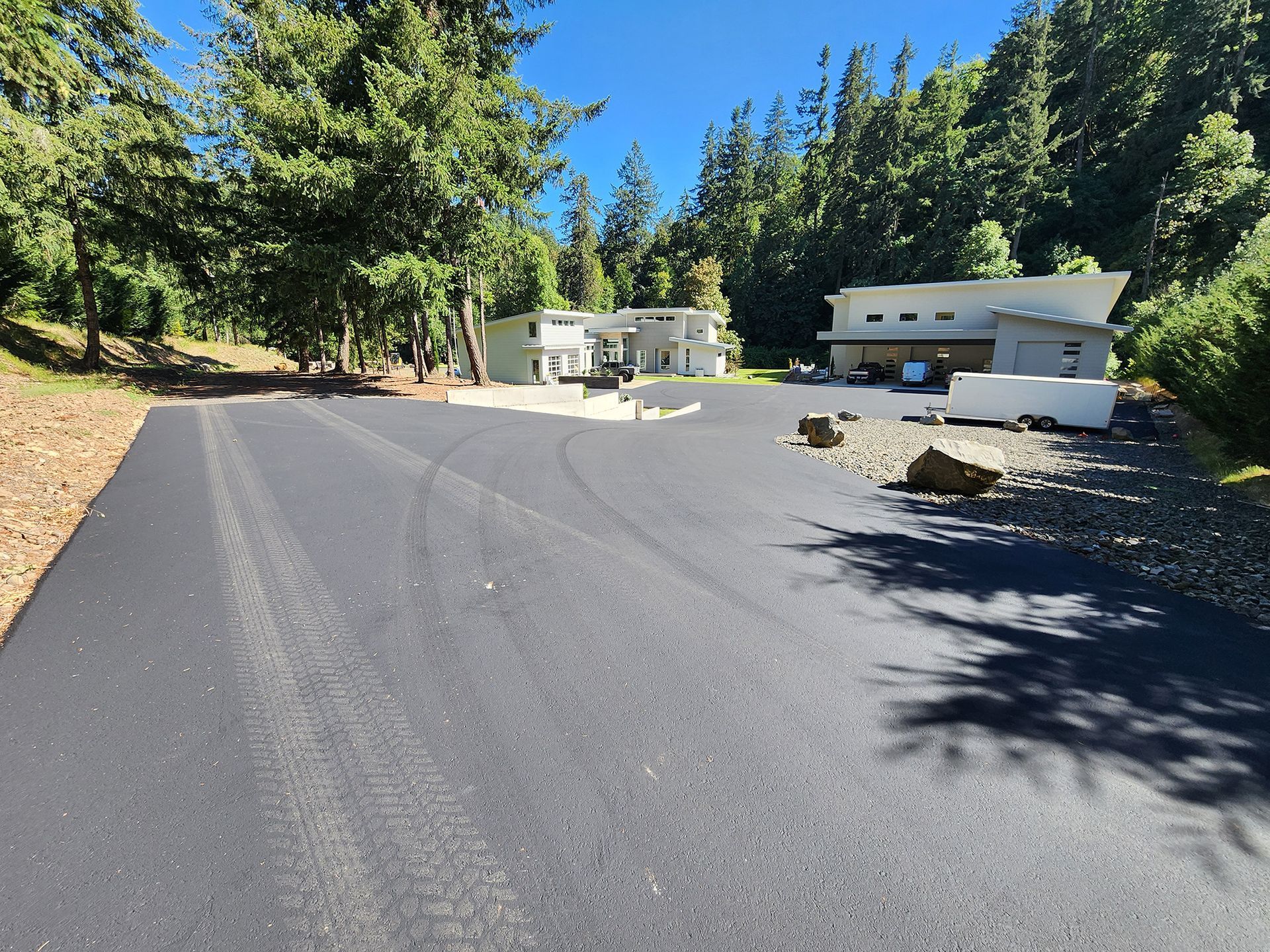 Paved driveway beside two white houses and tall pine trees on a sunny hillside