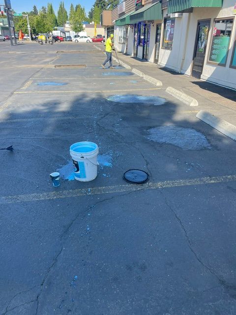 Parking lot with a spilled white liquid, blue bucket, and a worker in a yellow vest near storefronts.