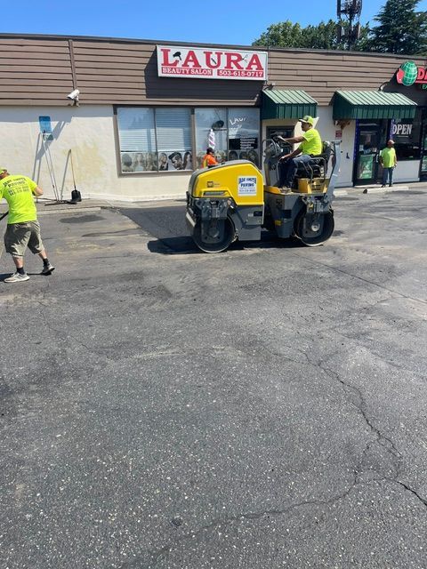 Workers paving a parking lot in front of a Laura store, with a yellow roller and one worker guiding it.