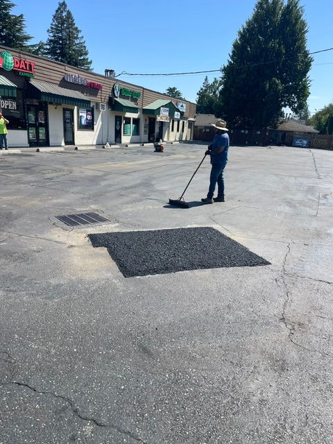 Person sweeping a parking lot beside storefronts with two dark asphalt patches on the pavement