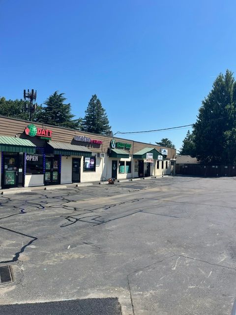 Small strip mall storefronts beside a cracked, empty parking lot under a clear blue sky.