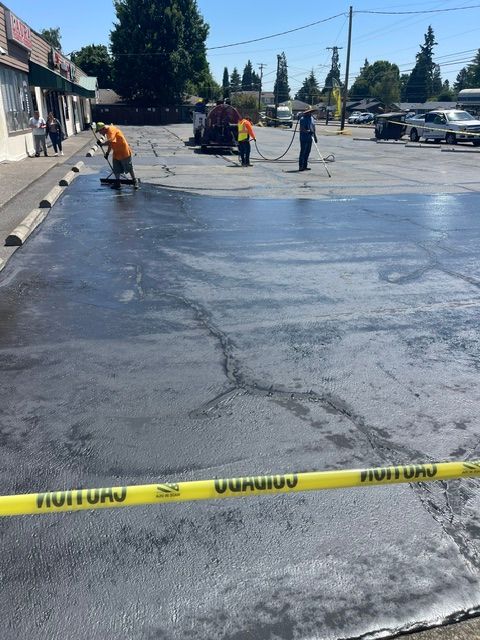 Road workers resurfacing a parking lot, with fresh black pavement and yellow caution tape in front.