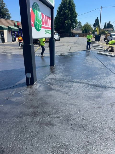 Workers resurfacing a storefront parking lot beside a black sign under clear blue skies