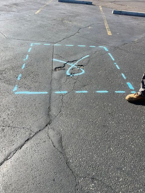 Blue dashed square painted on asphalt around a broken pavement feature in a parking lot; a shoe is visible at right.