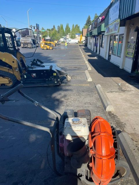 Construction machinery on a street beside shops, with a worker using a walk-behind machine.