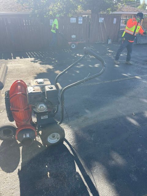 Worker in orange vest and face shield operating a hose by a red industrial fan on a sunny street.