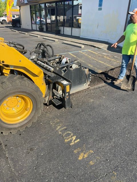 Yellow construction loader lifting a damaged curb section beside a storefront, with a worker standing nearby.