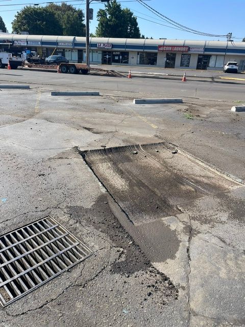 Damaged asphalt parking lot with a large pothole and storm drain near storefronts