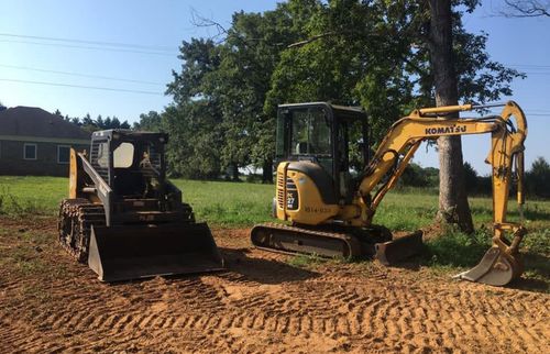 Yellow excavator beside a black skid steer on a dirt worksite near trees and a house