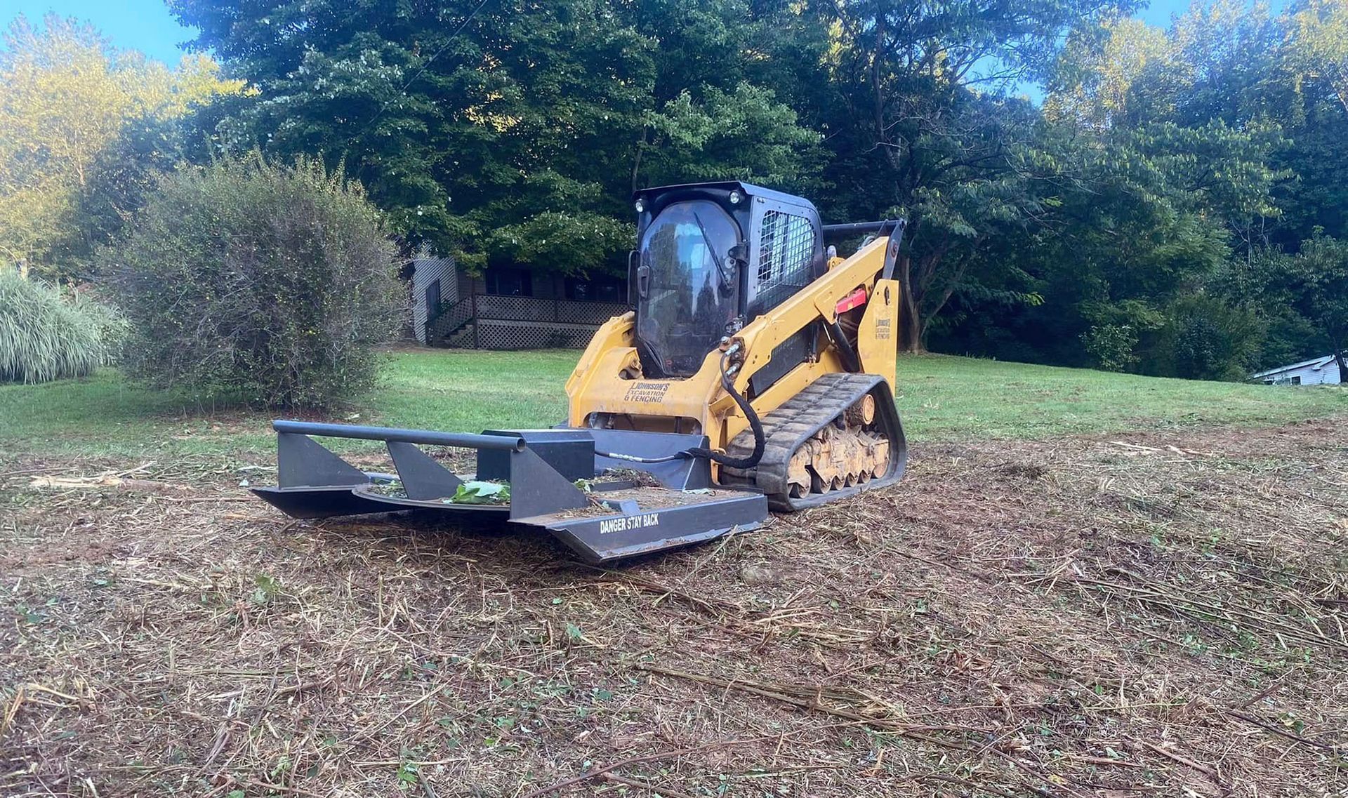 Yellow skid steer with front blade on a leaf-covered field, trees in the background