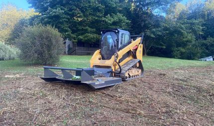 Yellow skid steer with front blade on a leaf-covered field, trees in the background