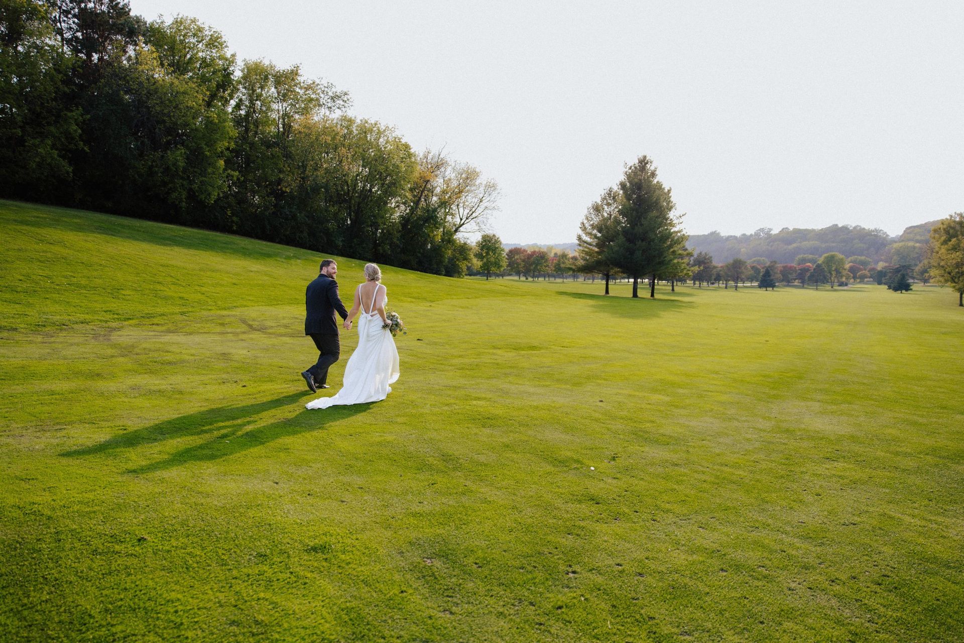Bride and groom walk across a sunny green lawn, trees and hills in background.