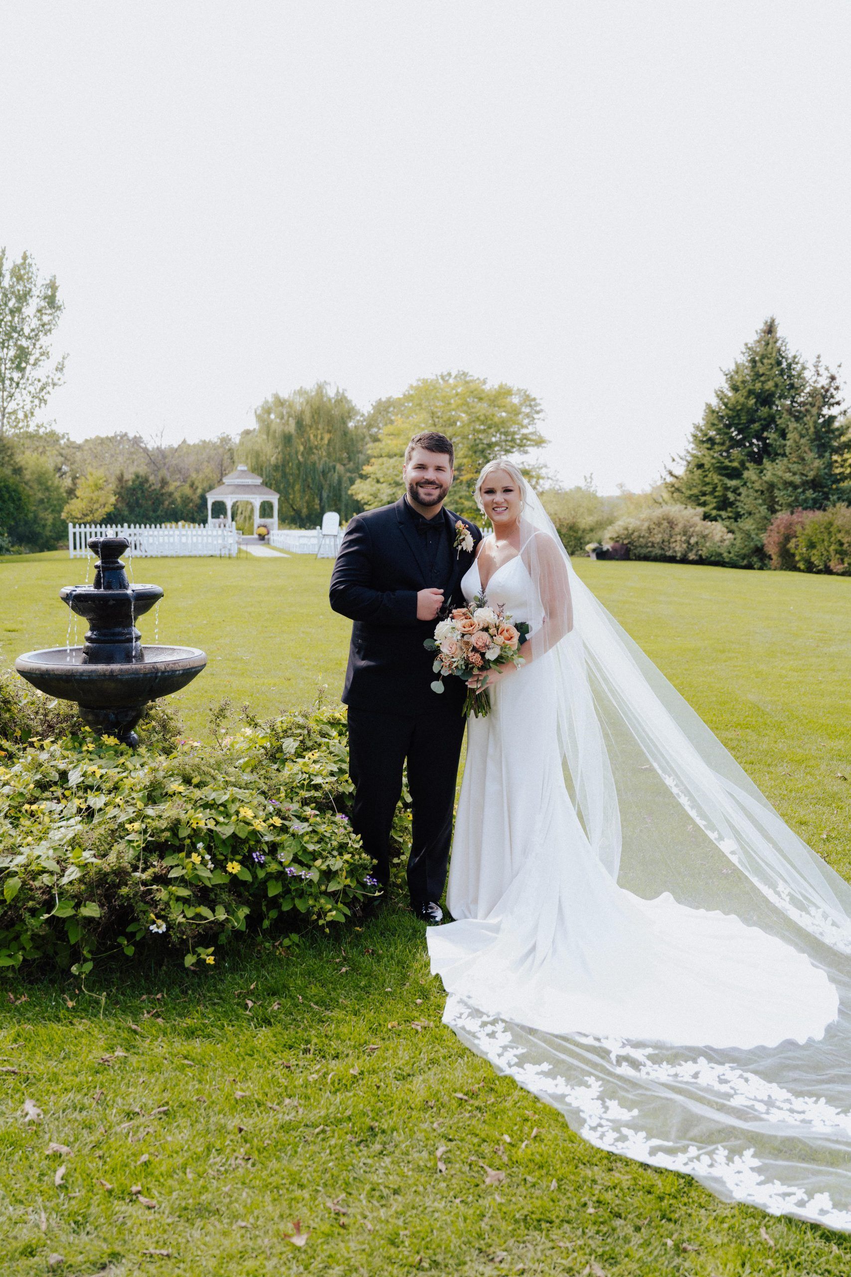 Bride and groom pose outdoors on their wedding day. Bride in white dress with long veil, holding flowers. Groom in black suit.