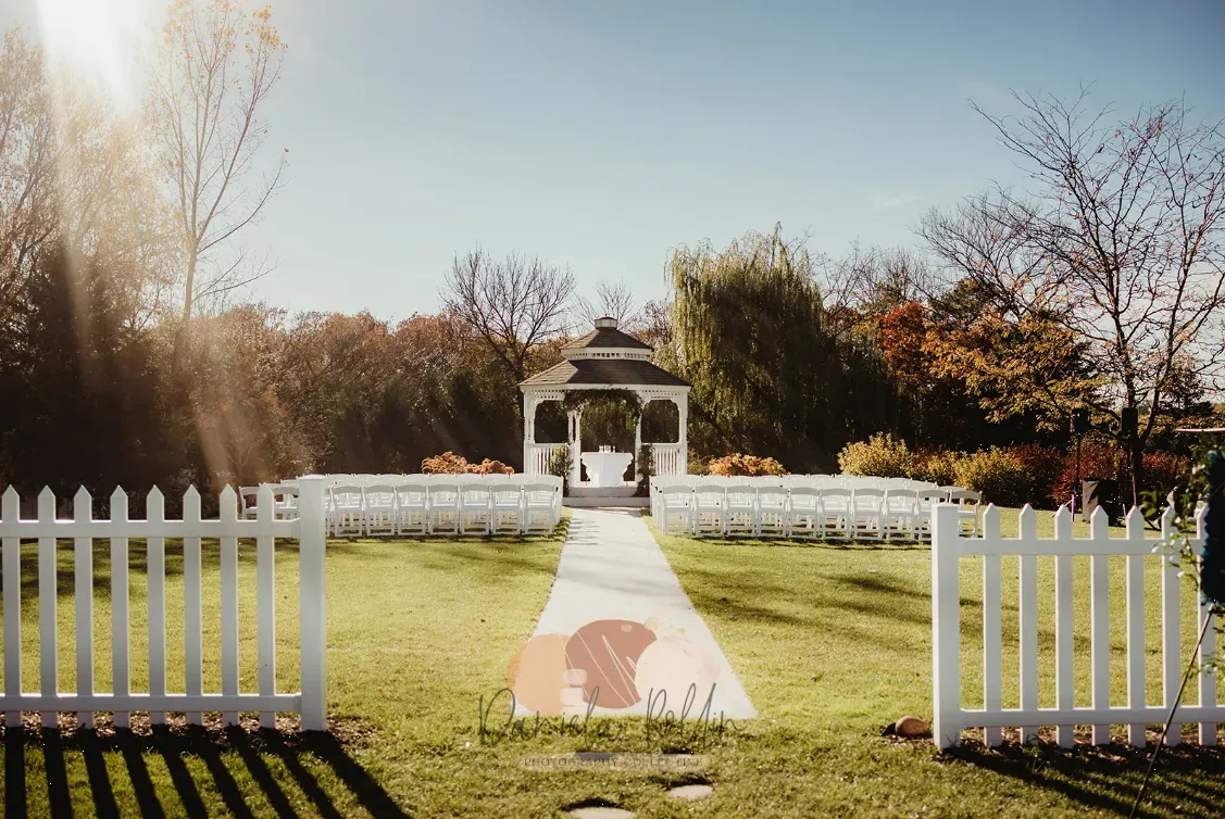 White picket fence leads to gazebo in a sunny garden, with autumn trees in the background.